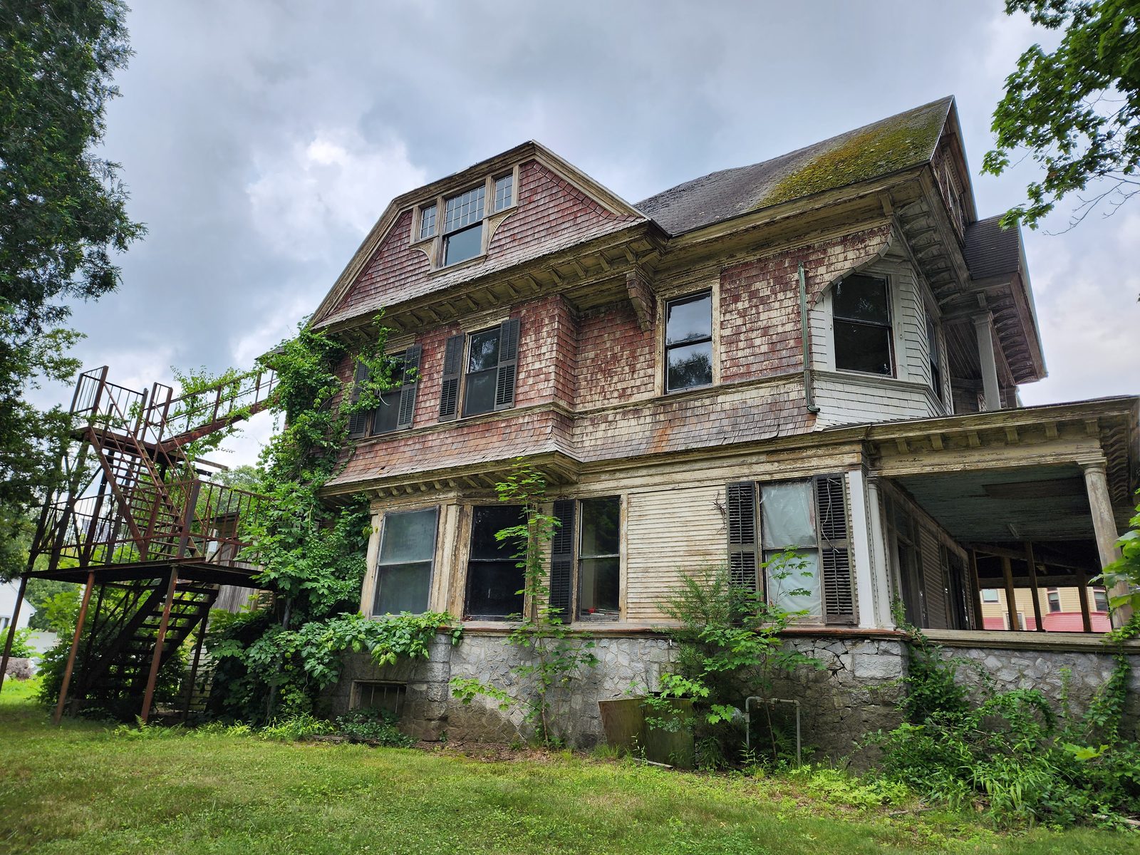 Left elevation before restoration: weathered red cedar shingle siding, a moss-covered roof, an exterior steel fire escape, shuttered and boarded windows, and vines climbing the stone foundation