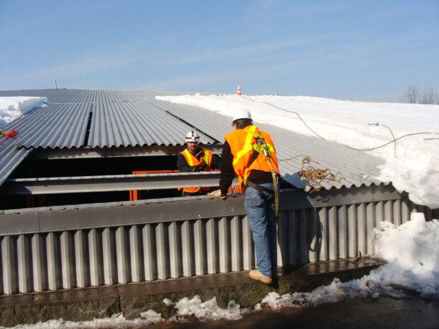 Workers in safety vests at an opening cut in the corrugated basin roof, with snow on the roof