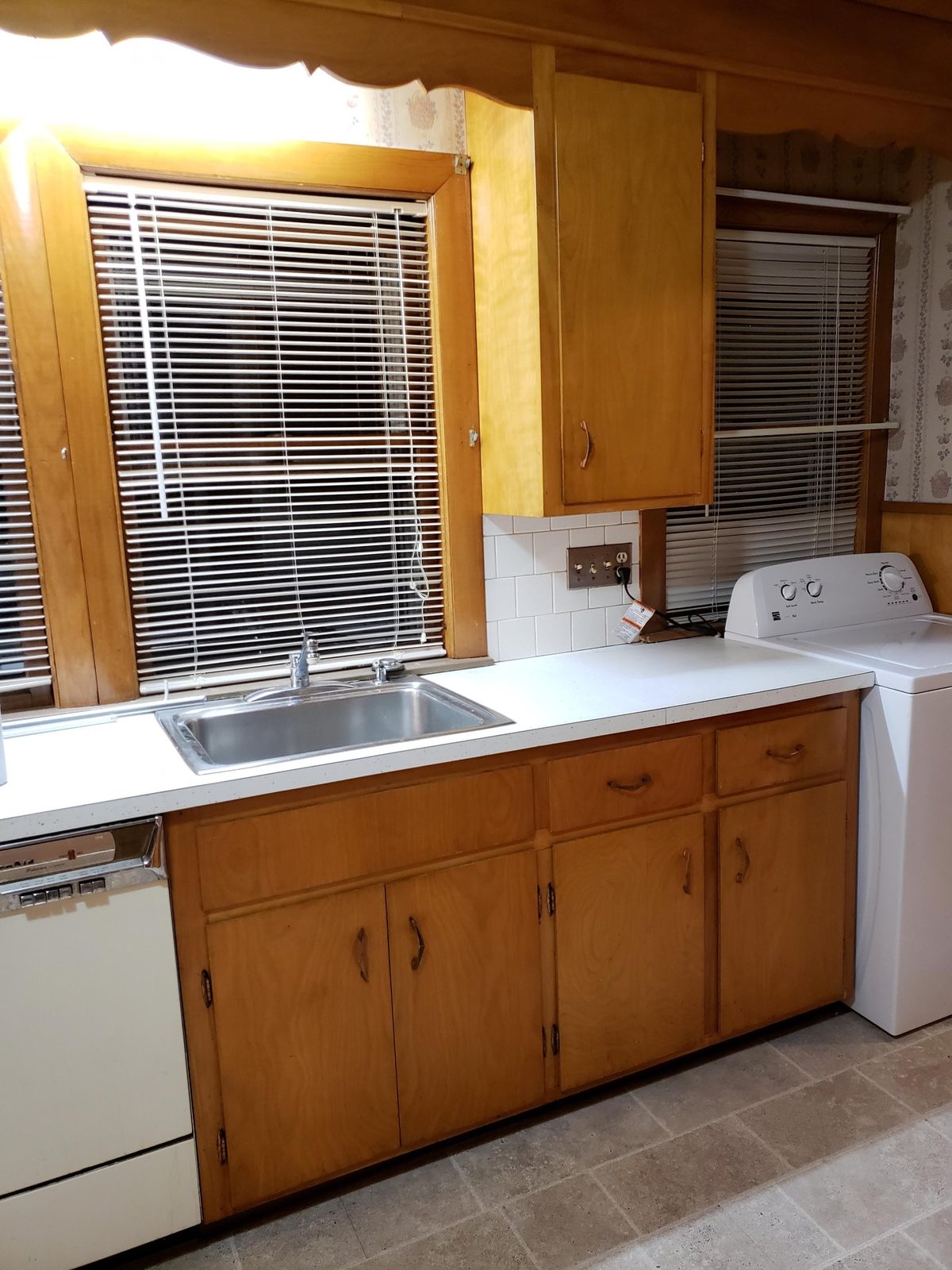 The original kitchen with wood cabinets, laminate counter, a top-load washer beside the sink and wallpapered walls